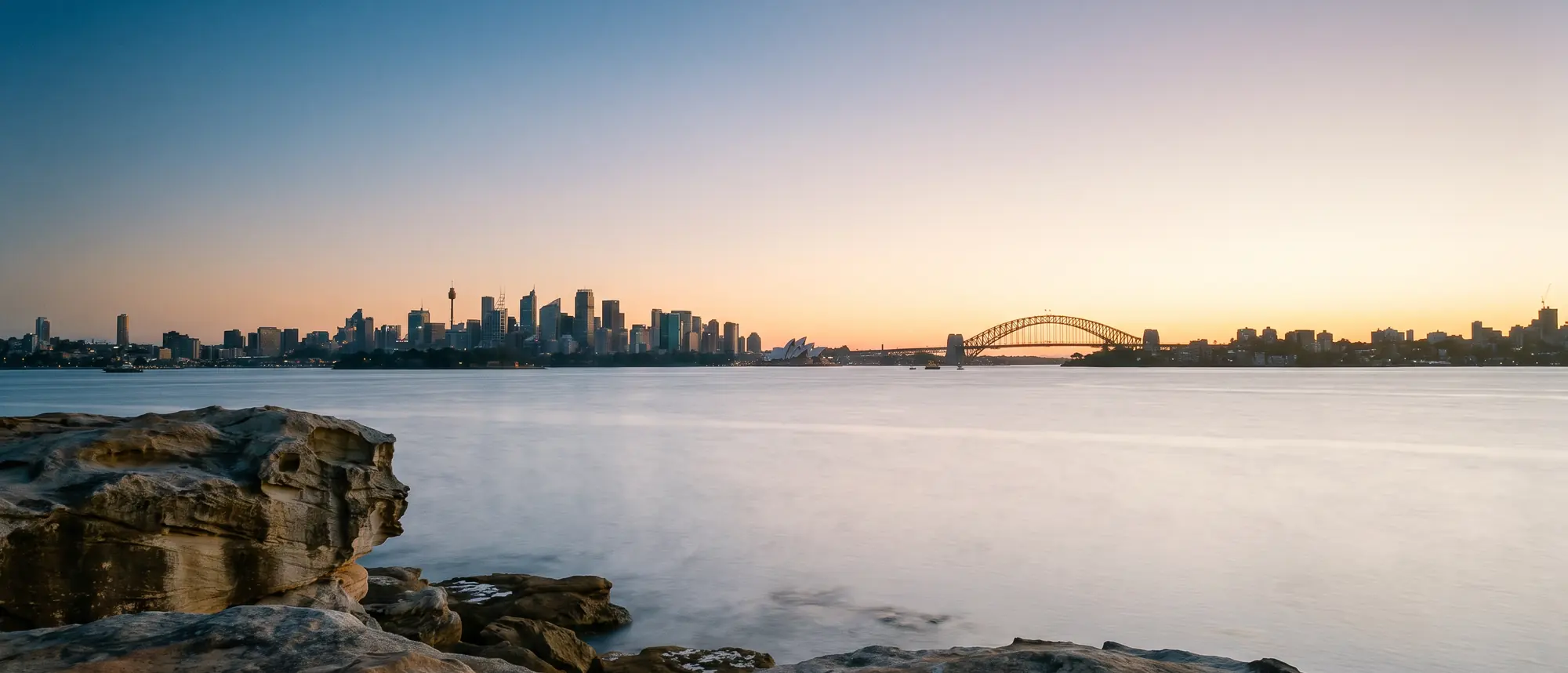 Sydney Harbour at first light from a sandstone headland, calm silver water under a wide gradient sky
